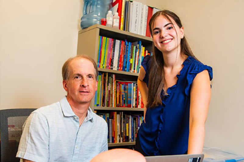 A man at his desk and a womans standing up next to him smile. A full book shelf is behind them