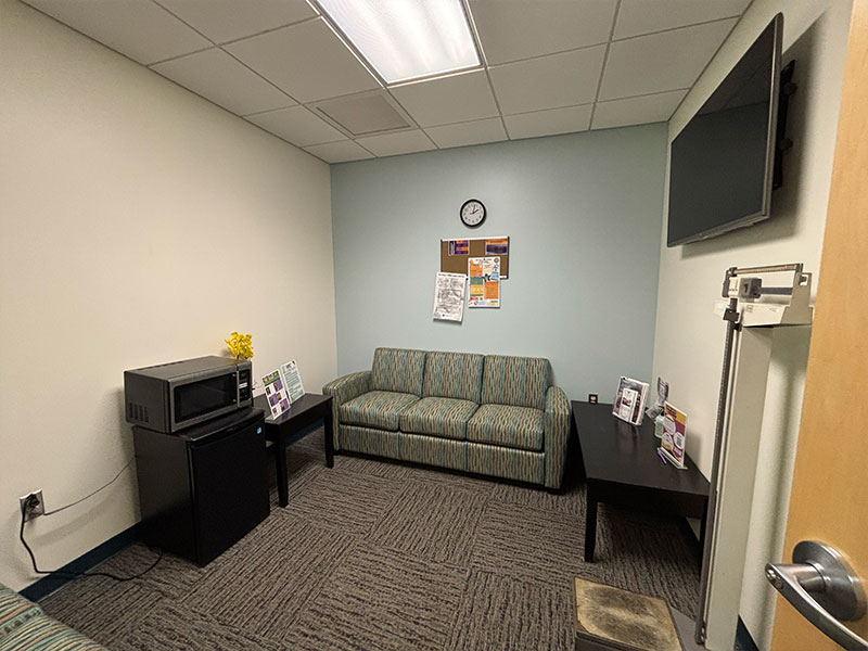 From door entry, on the back wall there is a large couch with side table. On the left wall, a mini fridge and a microwave. On the right wall, a longer table and a height and weight scale. 