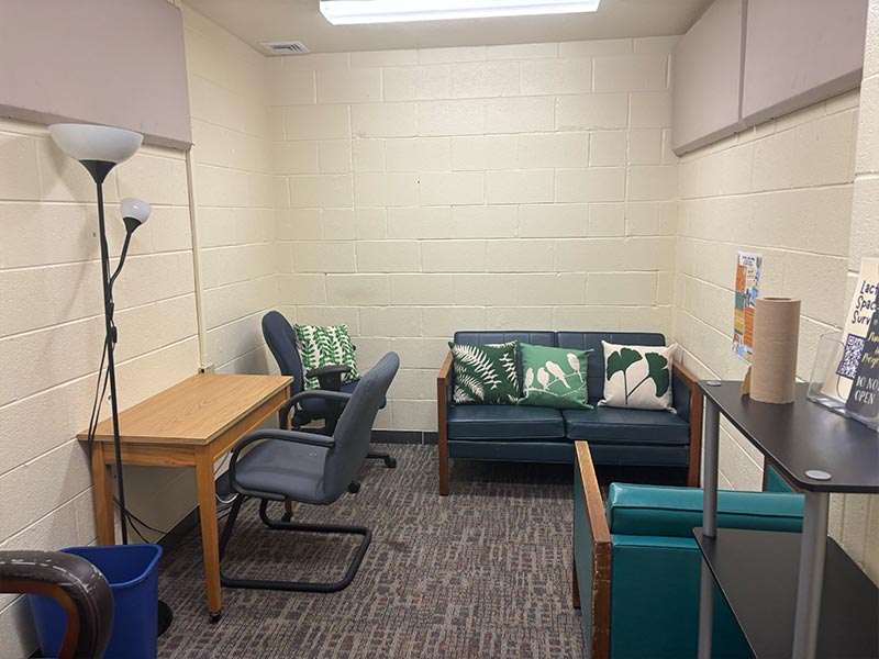 A room that includes a couch, an armchair, a desk with chair and a lamp. A table near the door has cleaning supplies. 