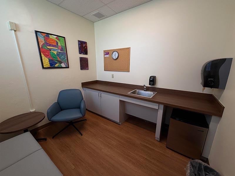 From the door entry, a couch is on the left wall, an armchair on the back wall with a side table in between and a sink, counter, and paper towel dispenser on the right wall. Under the right side of the counter, there is a silver mini-fridge.