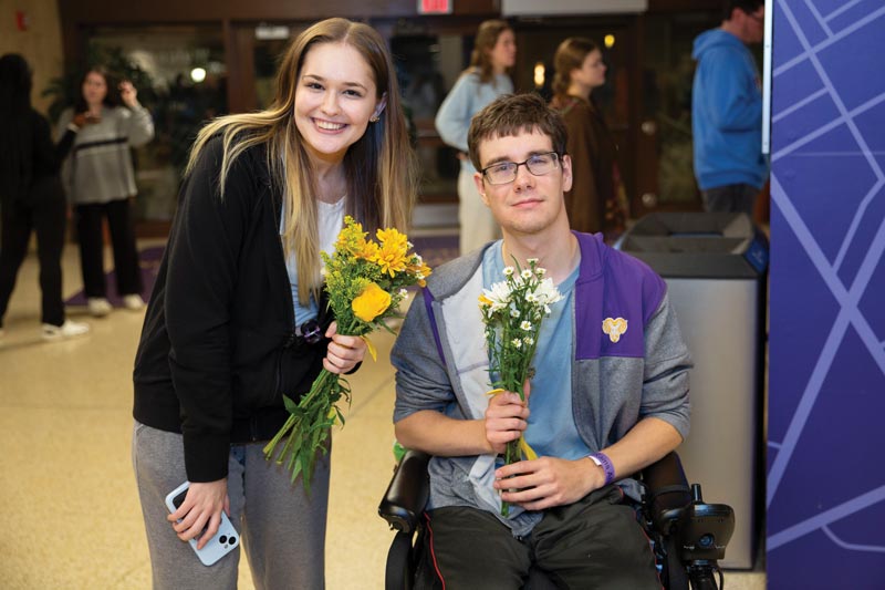 2 young people, one in a wheelchair, hold flowers and smile for the picture