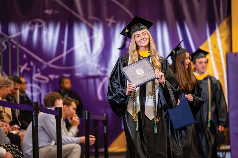 A student in her graduation robes holds her diploma and smiles