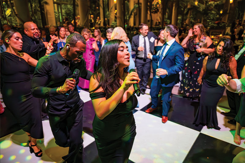 A woman and man each hold a microphone while dancing on a dancefloor surrounded by other people dancing