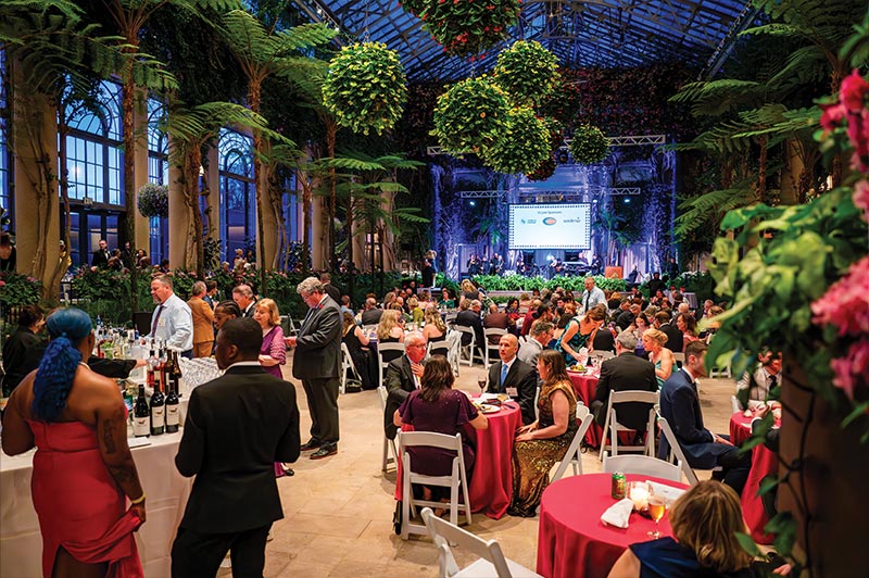 A room view of the gala featuring many people in fancy clothes sitting at tables, standing at the bar, and surrounded by flowers on the sides and ceiling