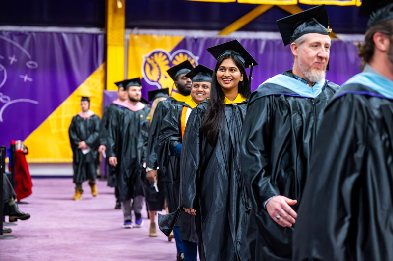 Graduates in black caps and gowns walk in a line during a graduation ceremony. A smiling woman is near the front, and purple and gold decorations are in the background.