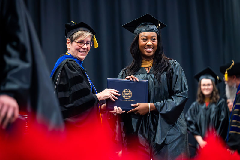 A graduate in a cap and gown smiles while receiving a diploma from a faculty member on stage during a graduation ceremony. Other graduates stand in the background.