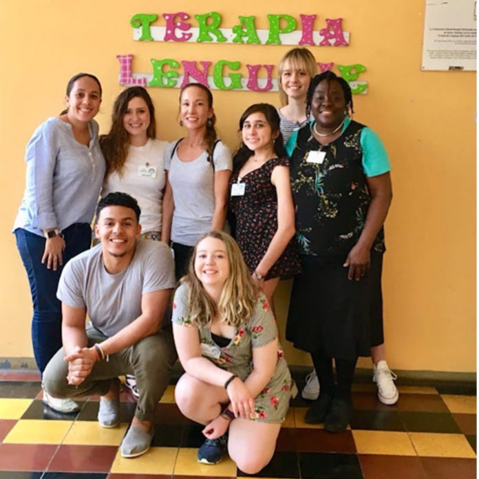 Eight women posing in front of a yellow wall. A sign reads Terapia Lenguaje.