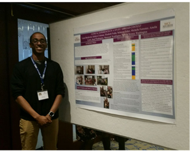 Student posing for camera. To his left is a poster.