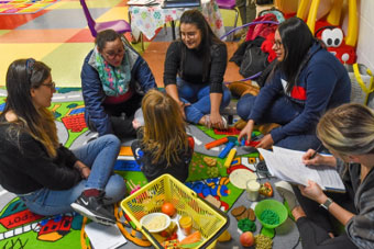 Classroom setting. 5 women sitting on the floor with a child.